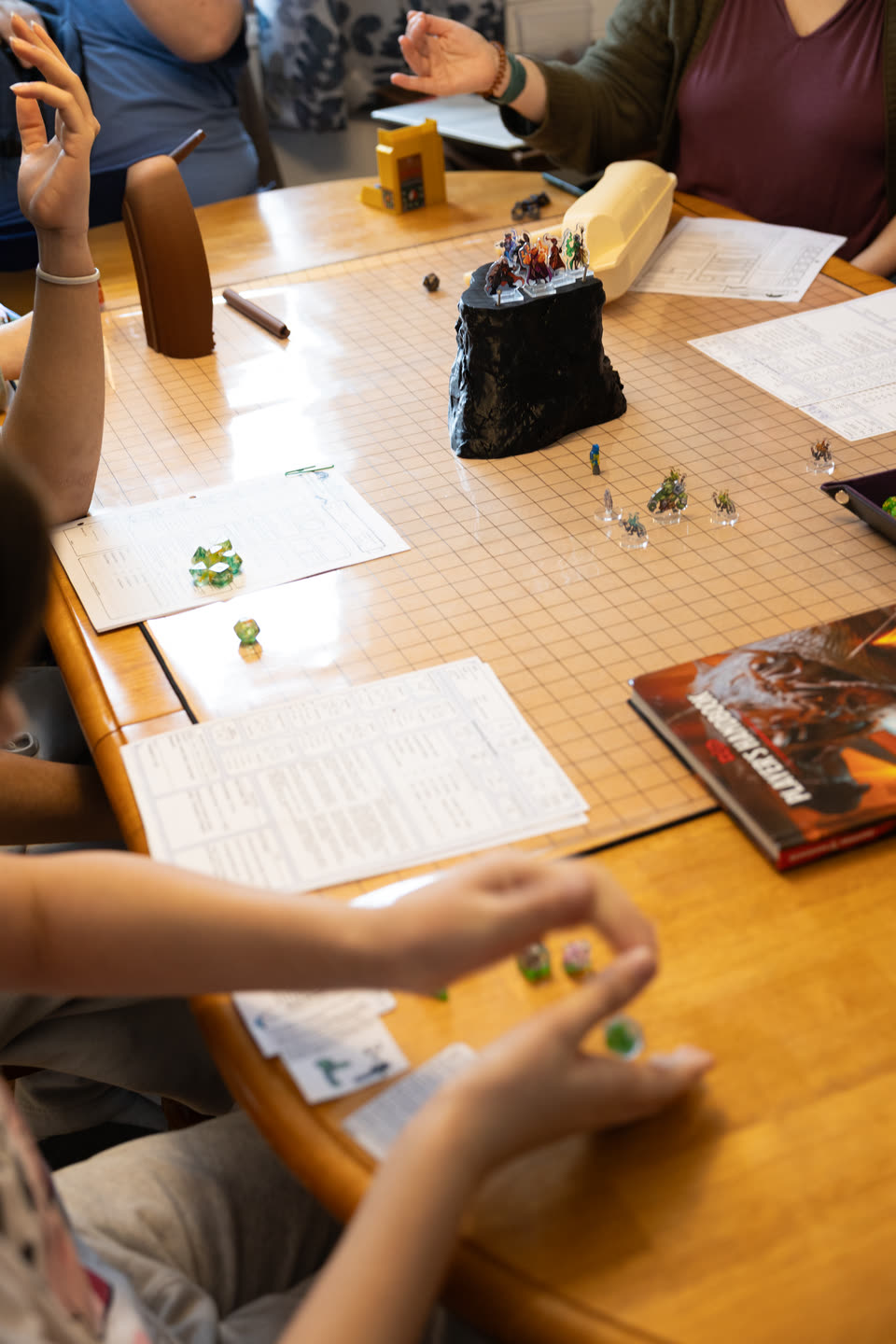 Players around the game table mid-turn: one raises a hand, another cups dice ready to roll, and miniatures stand on a rock tower in the middle of the map
