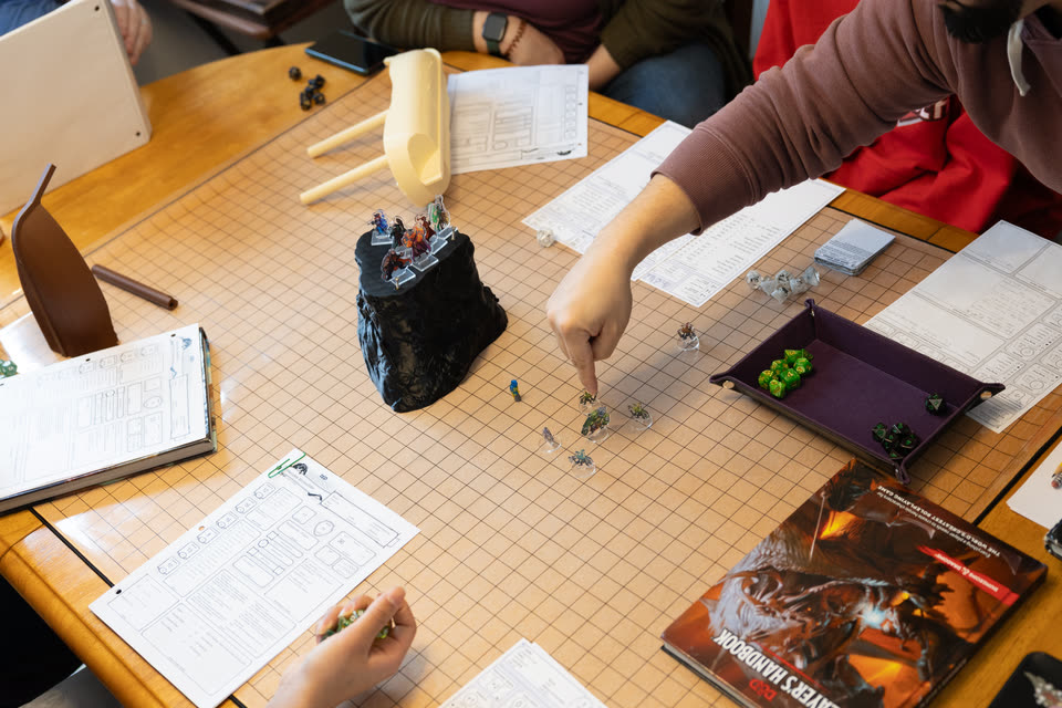 A player reaches across the grid mat to move a miniature beside a black rock tower, while others around the table hold dice and character sheets