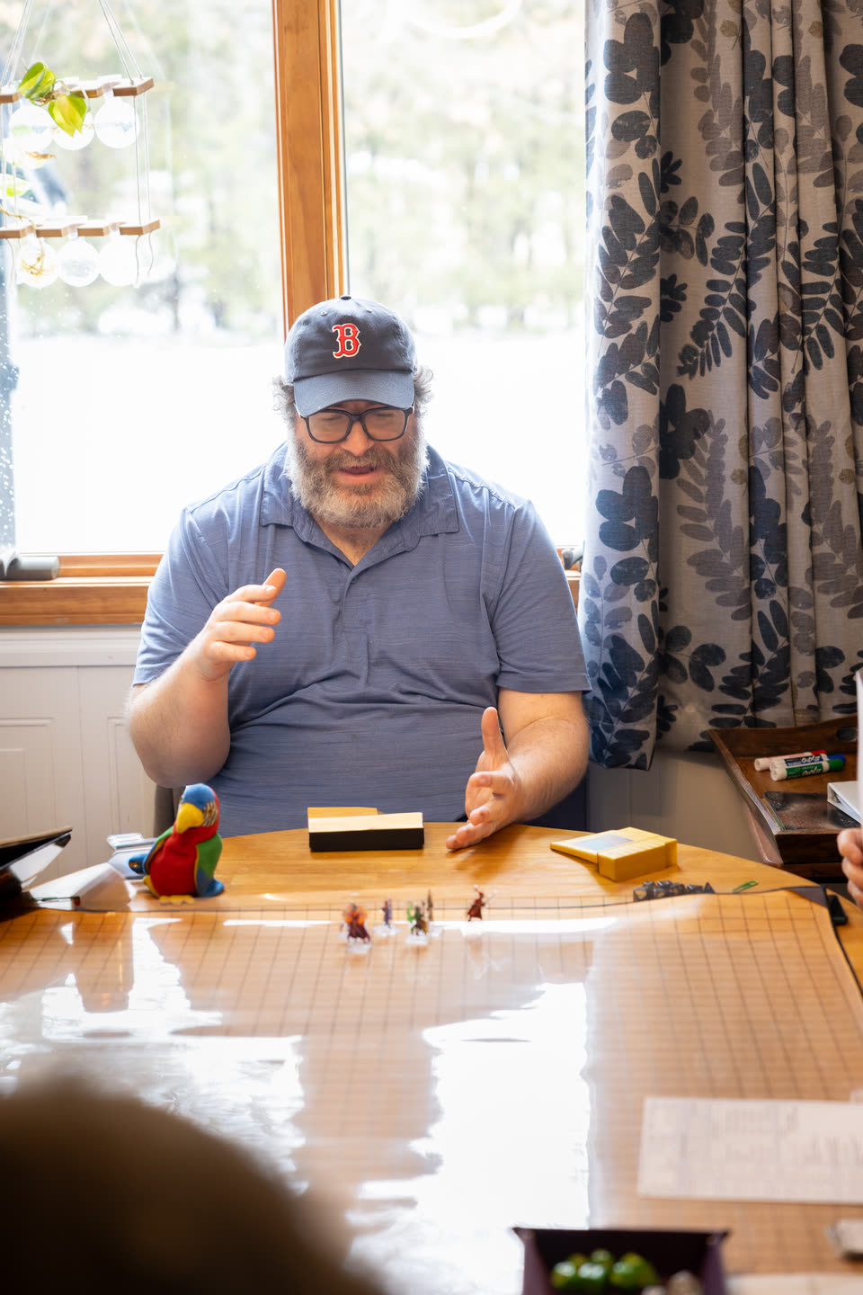 Steve, in a Red Sox cap, talks with both hands at the head of the game table, with painted miniatures on the grid mat in front of him