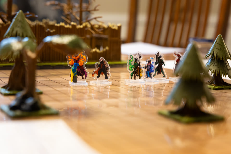 A party of hero miniatures stands among model pine trees, with a wooden fort behind them