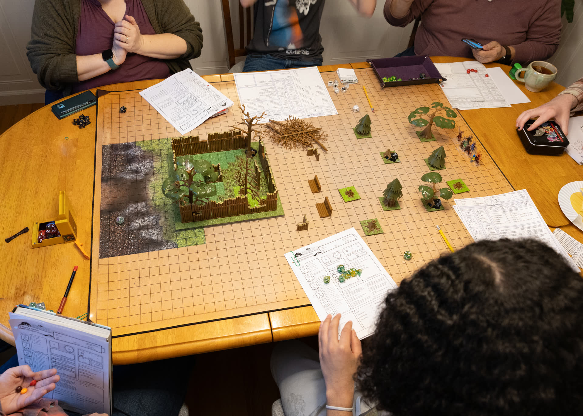 A group of adult players around a table set with a forest battle map, a walled fort, trees and miniatures