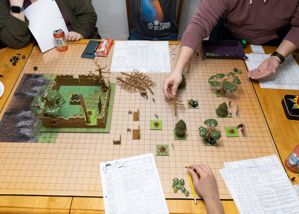 Adult players around a table set with a forest battle map and a walled fort: one moves a model tree, another holds out an open hand, and a third rests a pencil beside a character sheet